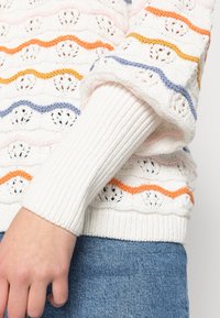 Strickpullover mit weißem Grund, der mehrfarbige Wellenstreifen in Orange, Pink, Blau und Gelb aufweist. Rippbündchen, strukturiertes Muster.