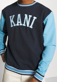 Navy blue sweatshirt with light blue sleeves, featuring the word "KANI" in bold embroidered letters. Ribbed cuffs and hem with striped accents.