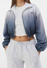 Giacca a vento cropped blu sfumato con polsini e vita elasticizzati. Presenta una zip frontale e un dettaglio ricamato con la scritta "Vale la pena aspettare".