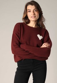 Pull en tricot bordeaux avec une texture douce, présentant un design de cœur blanc sur la poitrine gauche. Coupe ample et encolure côtelée. Associé à un jean noir.