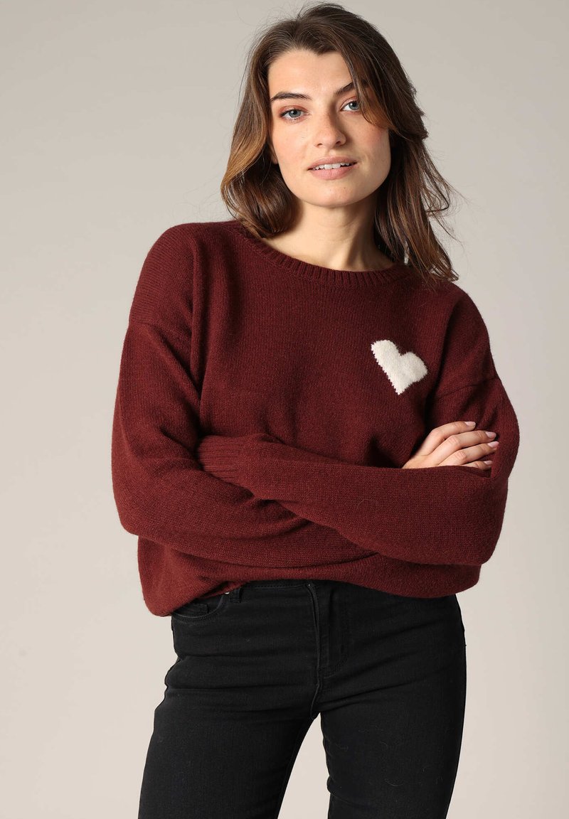Pull en tricot bordeaux avec une texture douce, présentant un design de cœur blanc sur la poitrine gauche. Coupe ample et encolure côtelée. Associé à un jean noir.