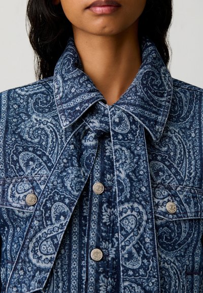 Donna che indossa una giacca blu con motivo paisley, con un ampio colletto e bottoni a pressione argentati, mostrata dal collo fino a metà petto.