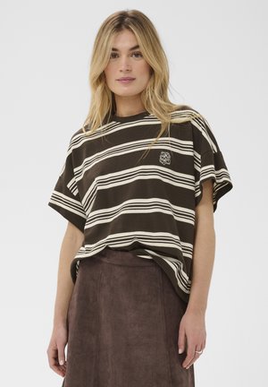 Femme blonde portant un t-shirt ample rayé noir et blanc et une jupe en daim marron taille haute, debout devant un fond uni.