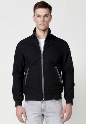 Chaqueta negra con cremallera y cuello alto, dos bolsillos frontales con cremallera, puños y dobladillo acanalados. Tela suave con detalles de diseño mínimos.