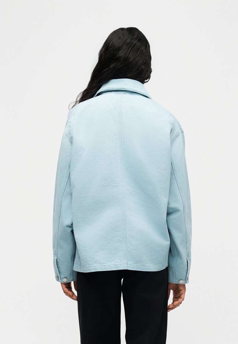 Person with long dark hair wears light blue jacket and black pants, facing away against plain white background.