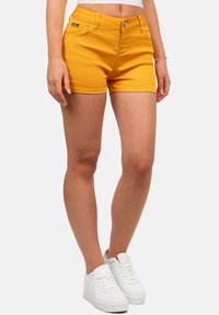 Gele denimshorts met een gladde textuur, vijf zakken en een knoopsluiting. Gecombineerd met witte sneakers.