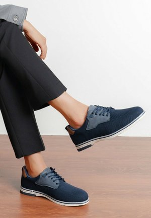 Personne portant un pantalon noir et des chaussures lacées bleu marine avec des semelles blanches, un pied levé, sur un sol en bois contre un mur blanc.