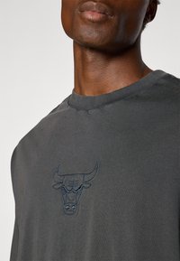 Mørkegrå t-skjorte med kort erm har en blå brodert okse-logo på brystet. Stoffet ser mykt ut med en glatt tekstur.
