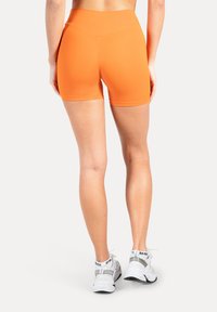 Orangefarbene Sportsshorts aus glattem, dehnbarem Stoff mit hohem Bündchen und figurbetontem Design, kombiniert mit weißen Turnschuhen.