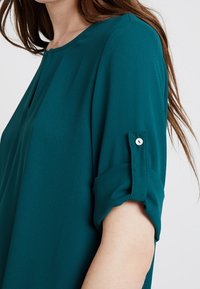 Miętowa bluzka z okrągłym dekoltem, krótkimi, podwiniętymi rękawami i detalem w postaci guzika na rękawie. Wykonana z lekkiej, miękkiej tkaniny.