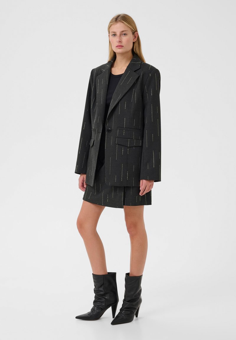 Blazer nero con motivo a righe verticali e due tasche frontali, abbinato a una gonna coordinata. Completato con stivali neri con punta a stringa.