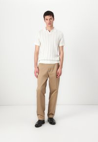 Pólo de malha branca com textura canelada, combinado com calças bege e calçado preto. Modelo em pé em um ambiente minimalista.