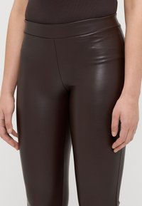 Leggings de pele sintética castanha com uma cintura alta e lisa, e um design ajustado e elegante. O acabamento da superfície é brilhante e refletivo.