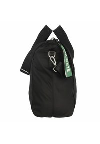 Sac de sport noir avec de larges bandoulières noires, clip en métal argenté, et étiquette en cuir vert Lacoste suspendue à la fermeture éclair latérale.