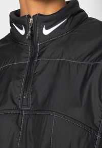 Czarny kurtka z zamkiem ćwierć, z białymi szwami i logo Nike na kołnierzu. Teksturowana tkanina z wysokim kołnierzem i zamkiem z przodu.