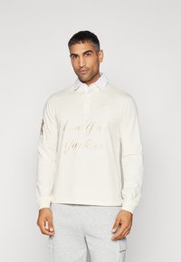 Cream long-sleeve polo shirt with white collar, featuring embroidered "New York Yankees" in gold. Material appears smooth and lightweight.