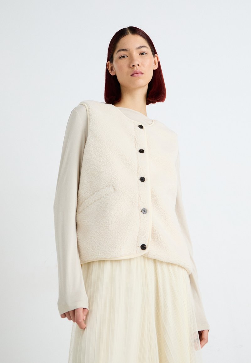 Krèmfarga fleece vest med rund hals, svarte knapper og sidelommer, lagd over en lys beige langermet skjorte.