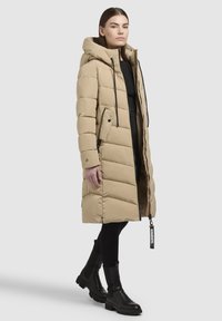 Manteau beige matelassé avec capuche, fermeture éclair à l'avant, poches latérales et accents en cordon noir. Assorti à des bottes noires à la cheville et à des leggings.
