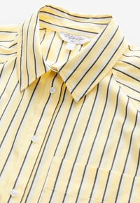 Chemise à boutons jaune avec rayures verticales noires et blanches, col pointu, poche avant et étiquette "The Girlfriend Shirt NEXT" à l'intérieur du col.