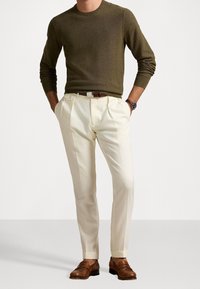 Olivgrön stickad tröja, krämfärgade veckade byxor, bruna läderskor och brunt bälte; klocka synlig på handleden. Enkel, skräddarsydd passform.