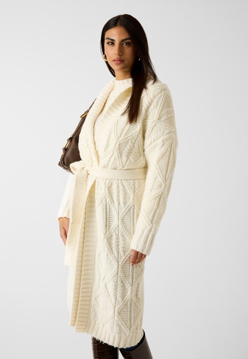 Gebreide crèmekleurige cardigan met een sjawlboord, gestructureerde patronen, een ceintuur in de taille en ribbelaccenten op de mouwen en de zoom.
