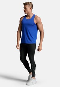 Blaues Sporttanktop kombiniert mit schwarzen Leggings mit texturiertem Design. Das Modell trägt weiße Sneakers. Schlichte, moderne Ästhetik.