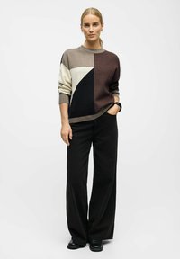 Trøjen har geometriske farveblokke i beige, brun, creme og sort med en rund halsudskæring. Den er parret med sorte højhalsede vide bukser.