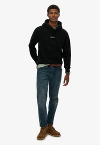 Sudadera de forro polar negra con bolsillo frontal y capucha con cordón, combinada con jeans de mezclilla azules y botas marrones con cordones. Camisa verde debajo.