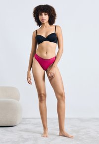 Czarny wzmocniony biustonosz i fuksjowy stringi Calvin Klein. Biustonosz ma cienkie ramiączka; stringi mają logo na pasie. Zestaw wyeksponowany na jasnym tle.