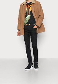 Giacca marrone con colletto chiaro su una camicia nera con motivo floreale, abbinata a jeans neri aderenti e sneakers Nike nere. Aspetto pulito e moderno.