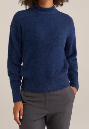 Persoon gekleed in een marineblauwe pluizige trui met een hoge hals en grijze op maat gemaakte broek, met één hand in de zak tegen een neutrale achtergrond.