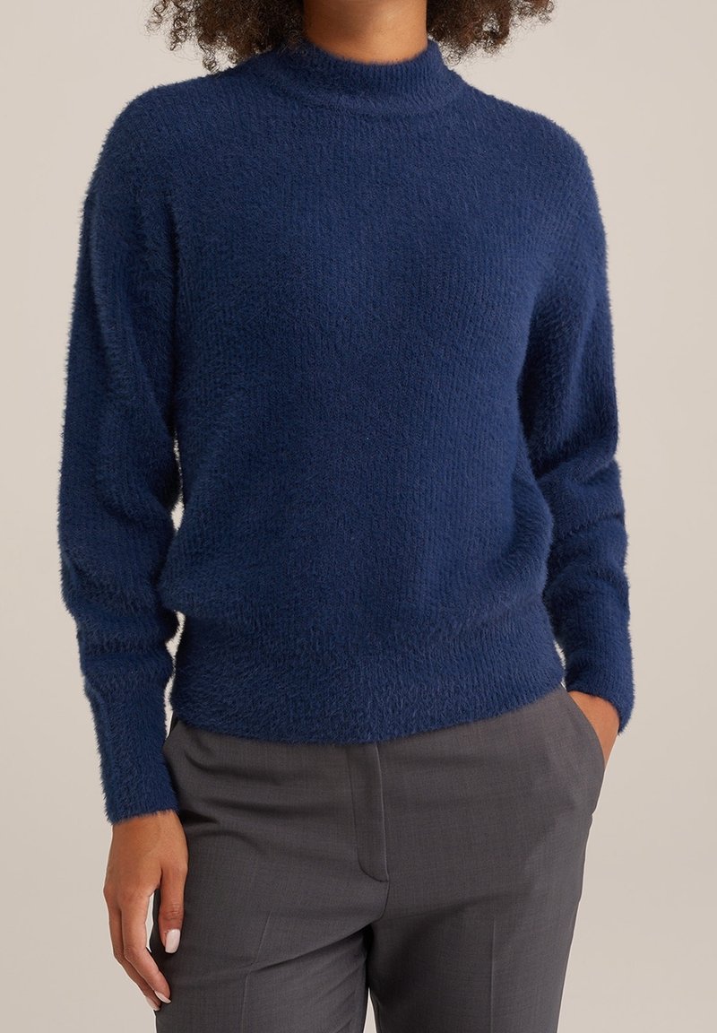 Persona che indossa un maglione morbido blu navy con colletto alto e pantaloni grigi su misura, con una mano in tasca contro uno sfondo neutro.