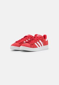 Pár červených tenisek Adidas Campus s bílými pruhy, bílými podrážkami a červenými tkaničkami na čistě bílém pozadí.