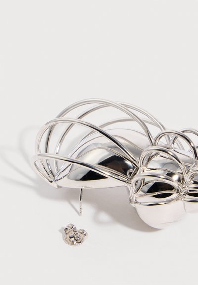 Ασημένιο μεταλλικό αφηρημένο μπροκάρ λουλουδιού με καμπύλα σύρματα σε σχήμα πετάλων και καρφίδα στο πίσω μέρος, πάνω σε λευκή επιφάνεια, δίπλα σε έναν μικρό μεταλλικό κούμπωμα.