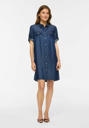 Femme portant une robe bleu foncé boutonnée à hauteur du genou avec manches courtes retroussées et talons ouverts marron, debout sur fond blanc.