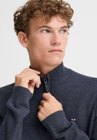 Suéter de punto gris oscuro con cuello alto y cierre de cremallera. Cuenta con puños acanalados y un pequeño logo en rojo, blanco y azul en el pecho.