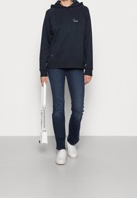 Persona che indossa un hoodie nero con un piccolo logo, jeans blu scuro, sneaker bianche e tiene una borsa bianca con tracolla a catena.
