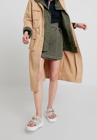 Abrigo largo beige sobre una camisa verde y una mini falda con cinturón. Sandalias de plataforma blancas con tiras. Texturas suaves y diseño en capas.