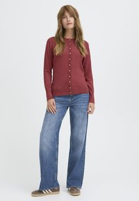 Roter Longsleeve-Cardigan mit goldenen Knöpfen, kombiniert mit hellblauen weiten Jeans und braunen Schuhen mit weißen Streifen. Minimalistisches Design.