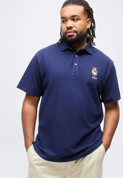 Άνδρας με ντέντλοκς που φοράει μπλε του ναυτικού πόλο με κεντημένη αρκούδα και μπεζ παντελόνι, με τα χέρια στις τσέπες, κοιτάζοντας προς τα πλάγια.