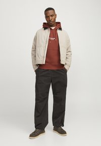 Giacca bomber beige sopra un hoodie bordeaux, pantaloni neri a vestibilità rilassata e stivali marroni con lacci e suole leggere. Tessuto morbido e design semplice.