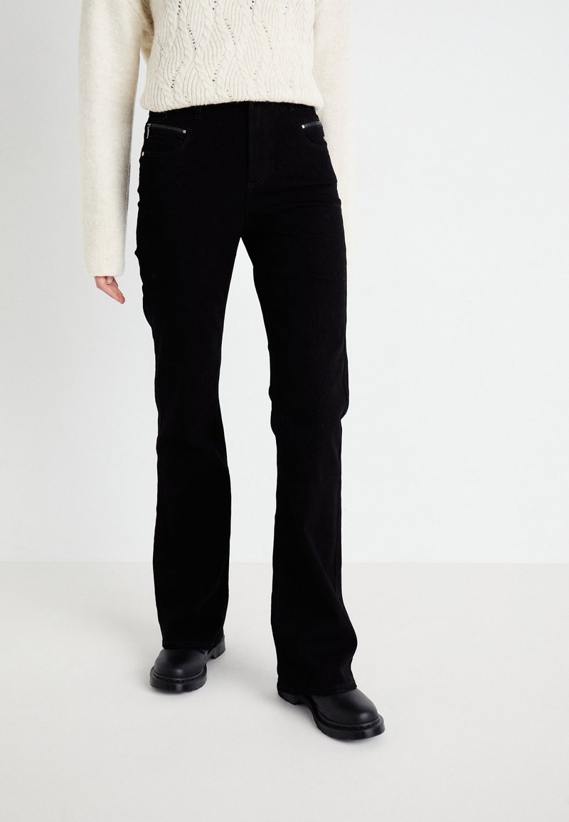 Pantalones acampanados de pana negra con bolsillos frontales con cremallera. Tejido texturizado y diseño de cintura alta. Combinados con un suéter de punto crema y botas negras.