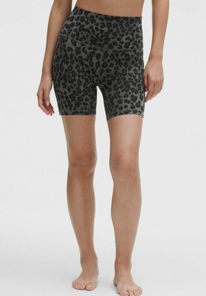 Frau trägt hoch taillierte Biker-Shorts mit schwarzem und grauem Leopardenmuster, steht barfuß vor einem schlichten, hellen Hintergrund.