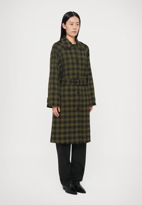 Pikk ruuduline mantel tumes rohelises ja kollases toonis, kraega kaeluse, nööpidega kinnituse ja vööga. Kandmine koos mustade pükste ja terava ninaga saabastega.