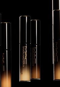 MAC Studio Radiance concealer-rör med svart och gradient brun ombre-design, med guldtext på en mörk bakgrund.