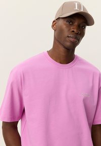 Ροζ βαμβακερό T-shirt με κοντά μανίκια, στρογγυλή λαιμόκοψη και διακριτική λεπτομέρεια λογοτύπου στο στήθος. Συνδυάζεται με μπεζ καπέλο με λευκές λεπτομέρειες.