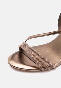 Κοντινή λήψη μεταλλικού χάλκινου σανδάλιου με ανοιχτή μύτη, τρεις λεπτές ραβδώσεις με χάντρες στο μπροστινό μέρος και λείο πέλμα.