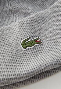 Grå ribbet strikhue med et grønt broderet krokodillelogo, der har en rød detalje på munden. Blød tekstur, afslappet design.