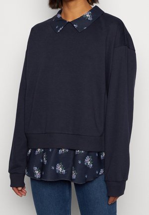 Person wearing a navy blue layered top with a floral collar and hem over blue jeans, against a plain white background.