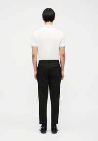 Άνδρας που στέκεται με την πλάτη προς το φακό, φορώντας λευκό πόλο με κοντά μανίκια, μαύρο παντελόνι καστορέ, μαύρες κάλτσες και μαύρα επίσημα παπούτσια.